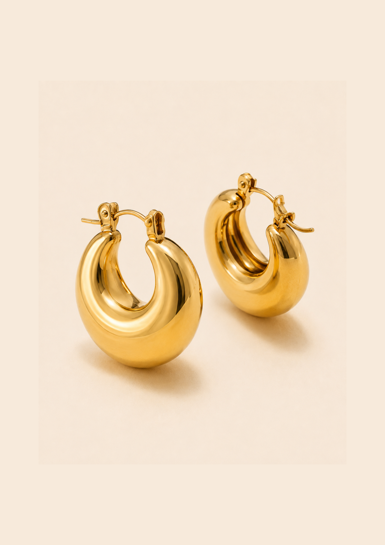 A pair of polished gold hoop earrings with a rounded, organic shape and visible spring hooks, displayed on a warm beige background.