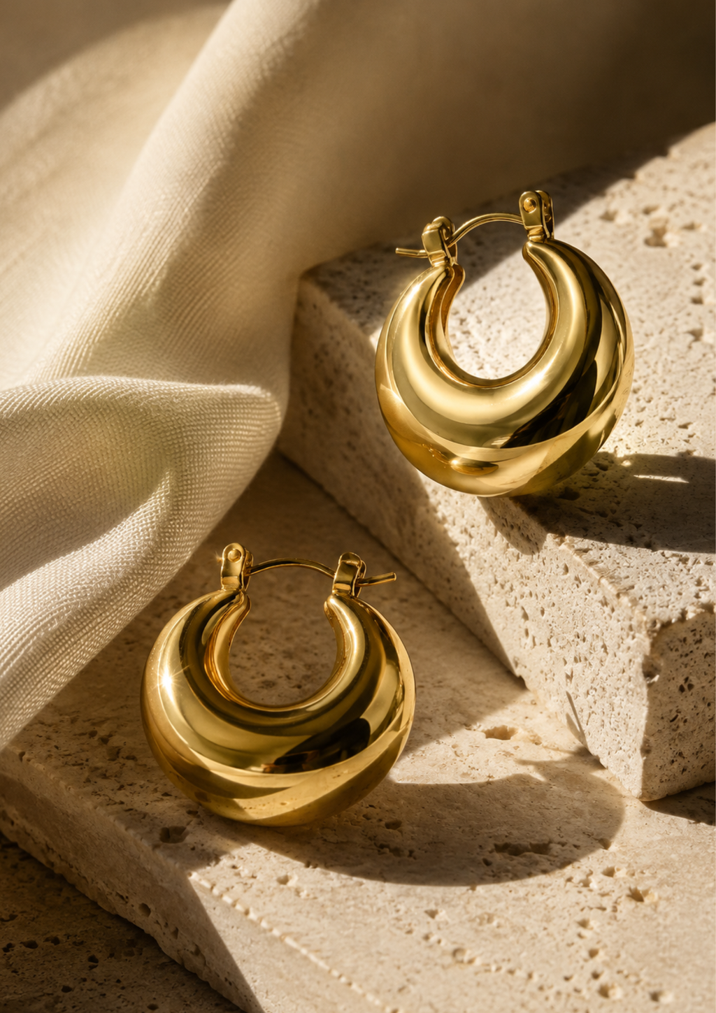 A pair of polished gold crescent-shaped hoop earrings displayed on textured stone blocks with draped cream fabric in warm, directional sunlight.
