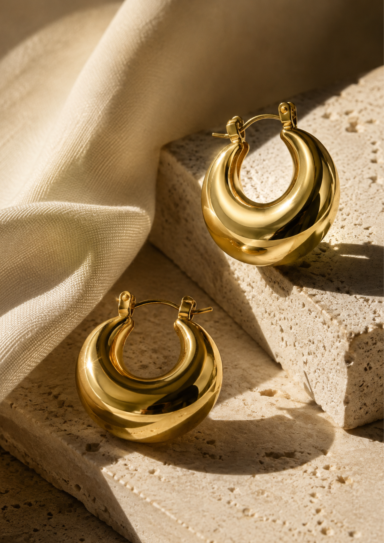 A pair of polished gold crescent-shaped hoop earrings displayed on textured stone blocks with draped cream fabric in warm, directional sunlight.